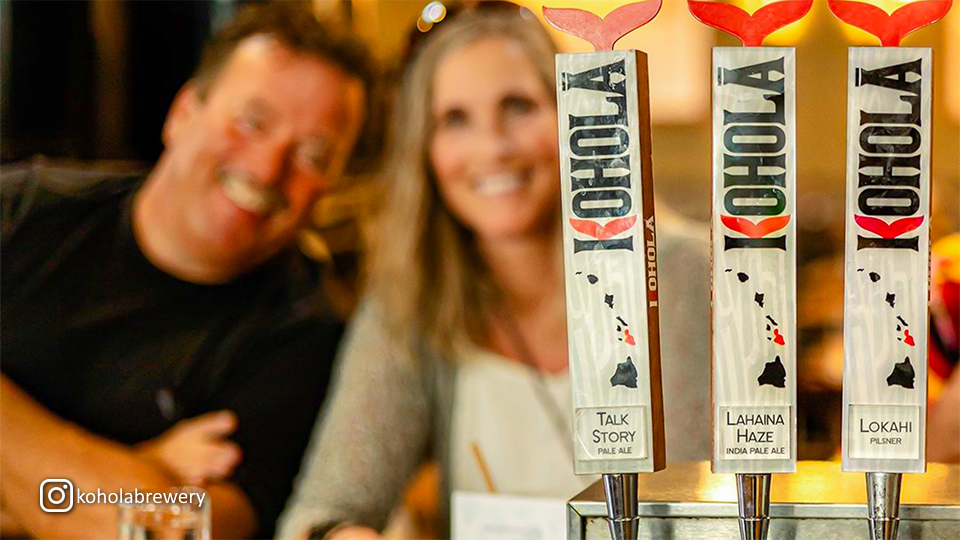 Close-up of three Kohola Brewery tap handles—Talk Story Pale Ale, Lahaina Haze IPA, and Lokahi Pilsner—with two people blurred in the background.