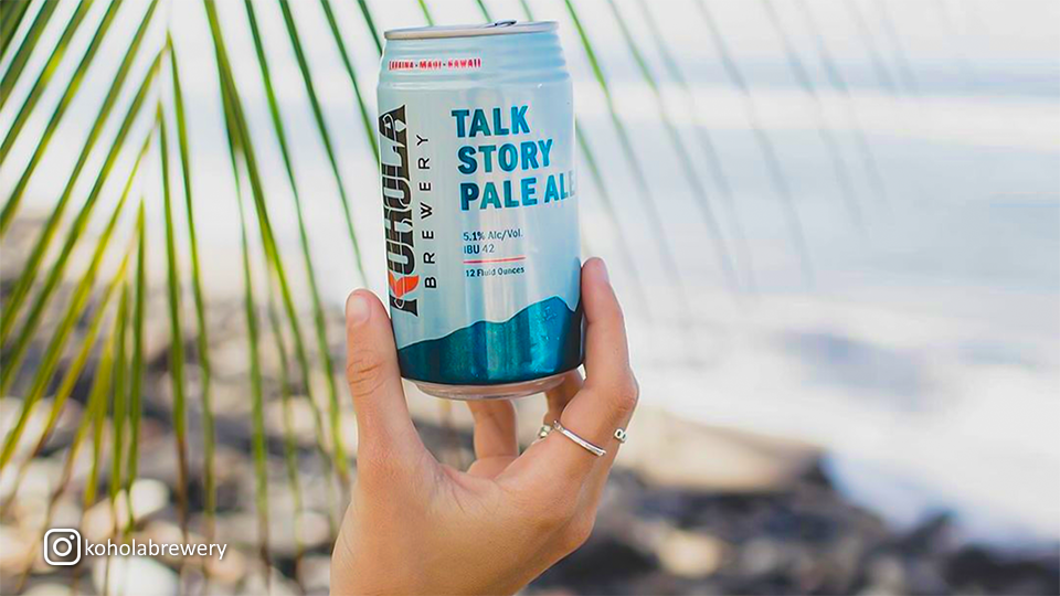 Hand holding a can of Kohola Brewery Talk Story Pale Ale with a beach view blurred in the background.