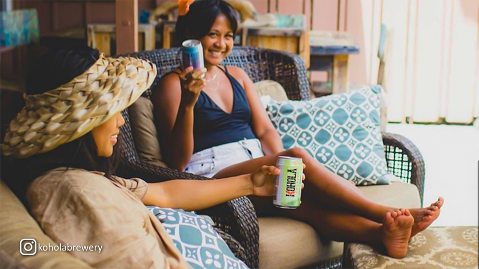 Two women lounging and smiling while holding cans of Kohola Brewery beer, enjoying a sunny day.