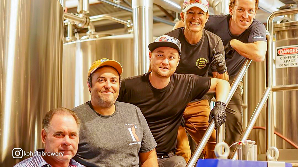 Group of five people standing with the brewhouse in the background at Kohola Brewery’s Tap Room, enjoying the brewery atmosphere.