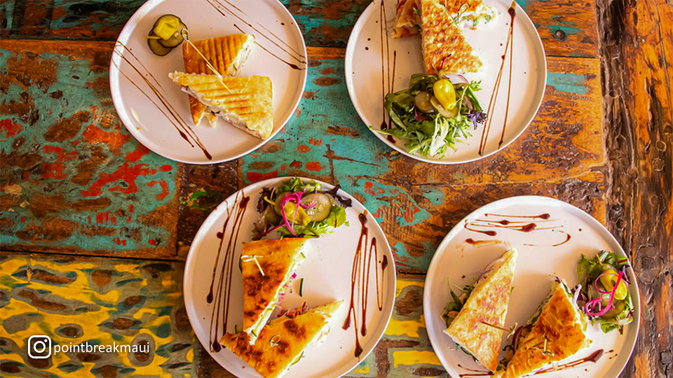 Overhead view of four different panini sandwiches at Point Break Pizza & Panini in Wailuku, Maui, each plated with unique ingredients and side dishes.