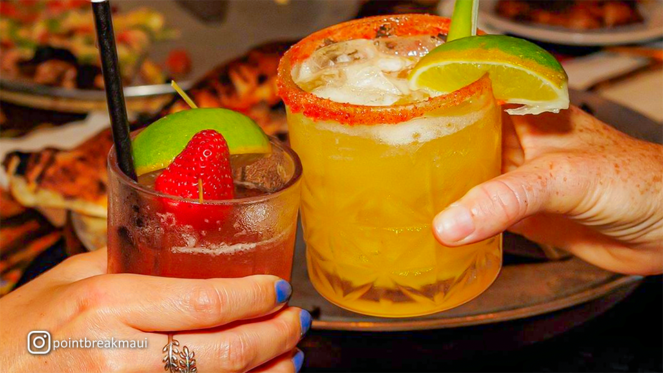 Two tropical drinks being cheered at Point Break Pizza & Panini, with colorful beverages served in glasses.